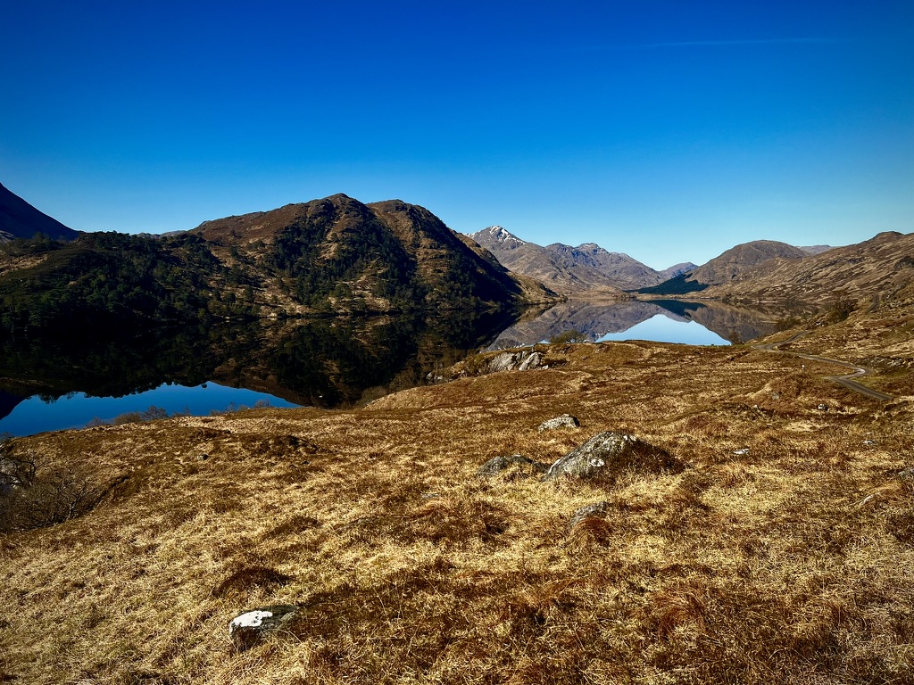 A Highland loch and surrounding hills reflected beneath a clear blue sky
