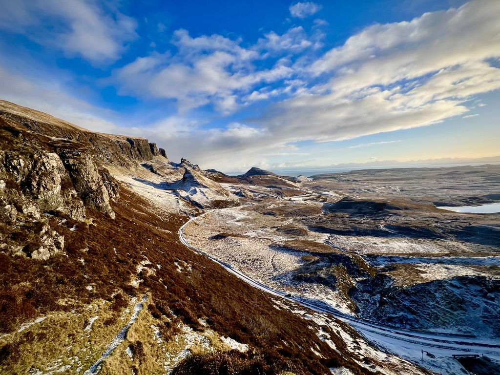 A winter Highland landscape of rocky escarpments, open moorland, distant water, and a winding road beneath a blue sky