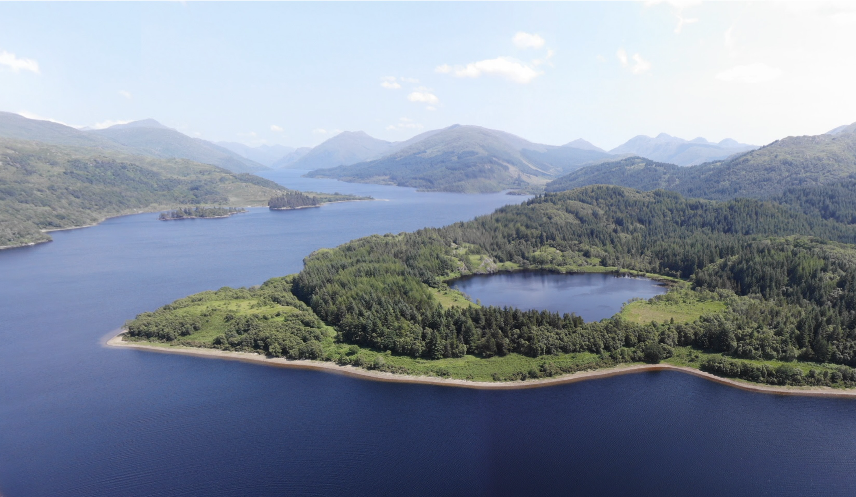 Finan’s Point extending into Loch Shiel, with its wooded peninsula, private lochan, surrounding water, and Highland mountains visible
